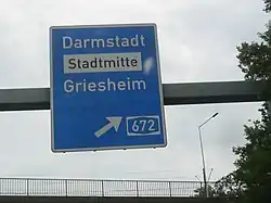 A sign on a German Autobahn: destination=Darmstadt;Stadtmitte;Griesheim destination:colour=;white;[2] destination:ref=A 672