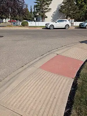 Red concrete tile at the sidewalk portion of a crossing that is flanked with concrete strips marked into the sidewalk parallel to the direction of travel. Across an asphalt street is a similar red crossing in concrete.