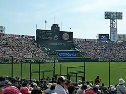 The outfield area of a baseball stadium with artificial turf, outdoors, seen from the stands near the fields; a fence roughly two meters high is atop the field wall. Beyond it, the rear wall has advertisements in Japanese with some brand names in English.