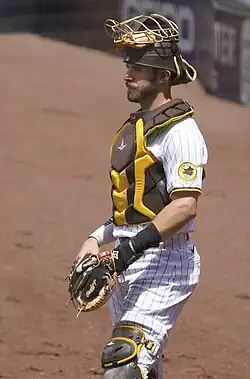 Austin Nola standing, wearing a pinstriped San Diego Padres jersey and brown and yellow catching equipment