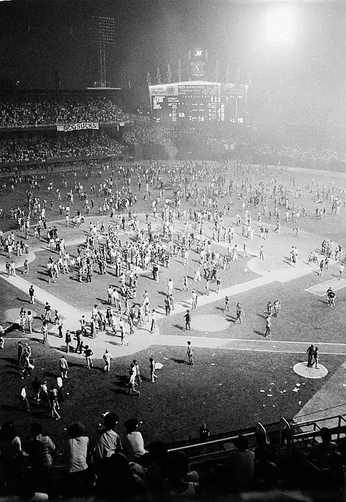 A color image of a crowd of young adults, mostly white men, walking around a baseball field and cheering. Some are holding signs, but what the signs say is unclear.