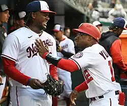 Eric Young Jr., wearing a red Washington Nationals helmet, glasses, and white uniform, smiling and putting his hand on the chest of Josh Bell of the Nationals
