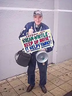 Full body shot of fan Freddy Sez, holding a pan with a shamrock and a sign that says "SCREAM-WHISTLE, KEEP UP NOISE!, IT HELPS YANKS!".