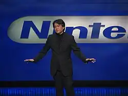 A Japanese man in a black business suit stands on stage with his arms spread outward some. Behind him is a blue wall with a blue Nintendo logo partially in view.
