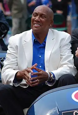A smiling, dark-skinned man wearing a white suit jacket, a blue button-down shirt and dark pants