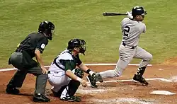 A baseball player in a gray uniform follows through with his swing, while a catcher on the opposing team in a white uniform squats behind him and an umpire in a black uniform stands behind the catcher.