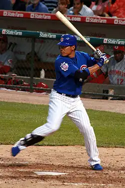 A man in a blue batting helmet with a "C" on it, blue baseball jersey, white pinstriped pants, and a shin-guard on his right shin stands at home plate holding a baseball bat in a left-handed batting stance with his right foot lifted off of the ground.