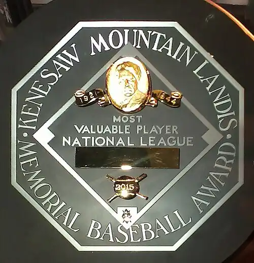 A black circle with an octagonal read "KENESAW MOUNTAIN LANDIS MEMORIAL BASEBALL AWARD". In the middle of the octagon is a baseball diamond which contains, from the top, Judge Landis' face in gold, "Most Valuable Player", the winner's league, his name in a gold rectangle, and his team.