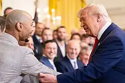 Mookie Betts in a tan suit, smiling and having his right arm gripped by Donald Trump at the White House