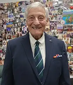 Elderly man with grey hair wearing a navy blue military blazer standing in front of a collage of photographs