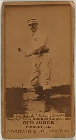 A man in a striped cap, white baseball uniform with the words "ST. LOUIS" obscured on the front and black belt pretends to swing an imaginary bat.