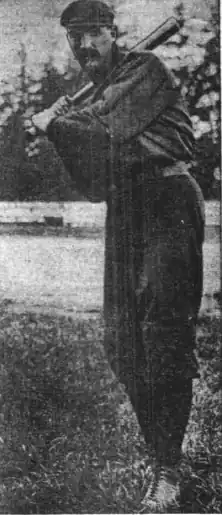 A man with a large mustache wearing a dark baseball uniform and cap stands poised to swing his bat.