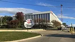 A large white building with multiple white columns and the lettering "WJW TELEVISION" at top center, against a blue sky and multiple trees in front of it. Right next to the building is a large white satellite dish with a "FOX 8" logo at top, a "CW55" logo in the middle, and the text "CLEVELAND'S OWN" at bottom.