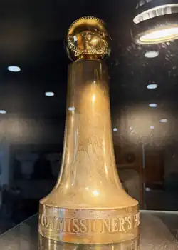 A photo of a golden trophy with a baseball on top of a tall, expanding cylindrical base with the letters "commissioner's h" visibly etched in the base