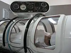 View of the side of a small cylindrical structure with several curved windows and an instrument panel, with a person visible inside