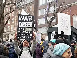 A placard saying that war only benefits the military industrial complex is held by a woman who smiles into the camera. Another protestor holds a peace symbol placard saying "Peace with Iran". Protestors are wearing winter clothing and the trees have no leaves. The background is filled with the walls of brick buildings.