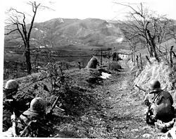 A machine gunner and two riflemen of Company "K" of the 87th Mountain Infantry Regiment, 10th Mountain Division, cover an assault squad routing Germans out of a building in the background. Porretta-Modena Highway, Sassomolare Area, Italy, March 4, 1945.