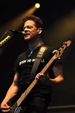 A middle-aged man wearing a black T-shirt holding a bass guitar and singing into a microphone