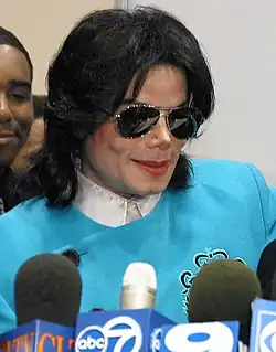 Jackson is wearing a white shirt and a light blue pullover. His skin is light and his nose is thin. He is wearing makeup and sunglasses. His hair is straight. Some microphones are standing on a lectern in front of him.