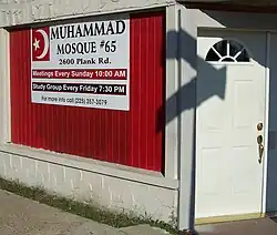 The front of a building. On the right is a closed white door; on the left is a window on which is a poster proclaiming "Muhammad Mosque Number 65".