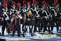 A group of performers onstage, with Madonna and Cee Lo Green at the front. They are all wearing black costumes with red and white stripes.