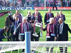 In the foreground is two men holding a large cup, they have claret scarves and a medal around their necks. Around them are ten old players in suits with medals and scarves around their necks