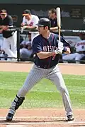 A man in a left-handed batting stance wearing pinstriped gray pants, a black shinguard on his right leg, a dark blue baseball jersey, and a dark-colored batting helmet.