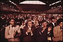 A large hall full of black men, dressed in suits and ties, clapping.