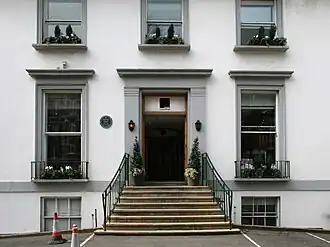 A flight of stone steps leads from an asphalt car park up to the main entrance of a white two-story building. The ground floor has two sash windows, the first floor has three shorter sash windows. Two more windows are visible at basement level. The decorative stonework around the doors and windows is painted grey.