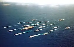 Aerial photograph of twenty-one ships, including an aircraft carrier, sailing in close formation.