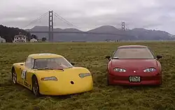 Two sports cars, the yellow one on the left, and a red one on the right, parked on a large, grassy plain. In the background is the Golden Gate Bridge.