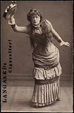 Reproduction of a sepiatone photograph of Adeleide Johannessen in corset and floor-length dress. She leans forward at the waist, her right arm stretched upward and outward, with a tambourine grasped in her hand. Her tense eyes look to her left. Written on one side of the card are the words Langaard's cigaretter! (which translates from Danish and Swedish as Langaard's cigarettes).