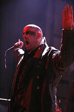 Head and upper body shot of a male singer, with a shaved head and blackened eyes, performing on stage. He is wearing a double-breasted military overcoat with a large collar.