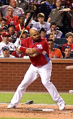 A right-handed batter is at the plate, looking toward the pitcher's mound. Wearing a red uniform and white pants, there is a crowd behind him with jerseys of various colors.