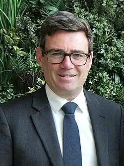 A fairly young man slightly smiling at the camera against green shrubbery as a background. He is wearing square glasses, a blue tie, and a grey blazer on a white shirt.