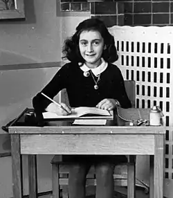 A black and white photograph of a teenage girl sitting at a wooden desk. She holds down the open page of a book with her left hand and has her right hand holding a pen over it, while smiling at the camera