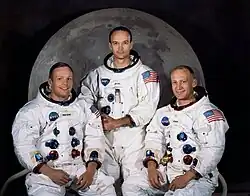 This official Apollo 11 crew portrait shows astronauts Neil Armstrong, Michael Collins, and Buzz Aldrin in their white NASA spacesuits, posed against softly lit backdrop of the Moon. Armstrong (left) and Aldrin (right) are seated, while Collins stands behind them at center. Each suit displays the astronaut’s name tag, the NASA insignia, and the American flag on the sleeve, with colored connectors visible on the chest.