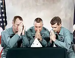 A color photograph of three seated men in a light green overall uniform, praying with folded hands towards a model of a spacecraft