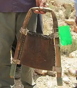 The hand of a man carrying a hoop made from a plant stem framing a leather bag, connected on the right side to the lower part of a green plastic bottle.