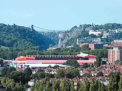 In the foreground, twentieth-century housing can be seen amidst trees, and on the right, a tower block of flats. In the middle distance, a complex of red coloured buildings can be seen, and behind that, a steep-sided gorge with a suspension bridge spanning it. Eighteenth-century terraces on the right side of the gorge, the slopes of which are heavily wooded, and a tower can be seen in the distance on the skyline.