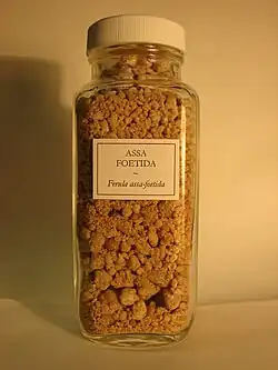 Pieces of resin of varying sizes and brownish-pink color in a vial labeled "Assa foetida – Ferula assa-foetida".
