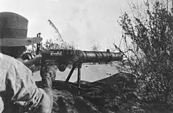 A soldier lies among the grass in the prone position behind a machine gun which he is holding in the shoulder.