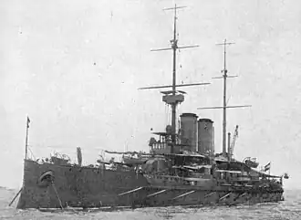 A large battleship at anchor in calm waters with two tall masts and two round funnels.