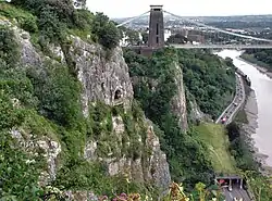 Rocky side to a gorge with a platform in front of a cave halfway up. To the right is a road and a river. In the distance are a suspension bridge and buildings.
