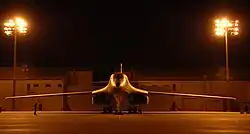 Front view of B-1 parked on ramp at night. Nearby yellow flood lights illuminate the area. In the background are buildings