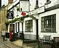 White fronted buildings with windows with small panes of glass. Shop signs for fish and chips and a pub. Postbox on the pavement in front of the buildings separated by black railings.
