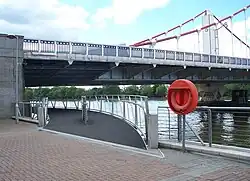 Start of a footbridge that curves under one end of a suspension bridge, along the bank of a wide river.