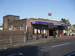 A red-bricked building with five people standing in front of it and a blue sign reading "BECONTREE STATION" all under a clear, blue sky