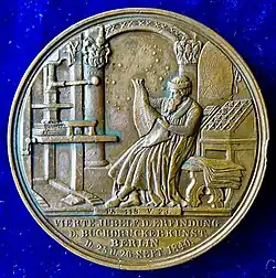 The reverse of the medal: Johannes Gutenberg in his workshop, sitting in front of his printing press