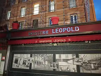 Exterior view of Brasserie Léopold in Aix-en-Provence, showing red signage, awning, and vintage photographs displayed on the storefront.